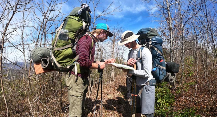 Two people wearing backpacks examine a map in a wooded area